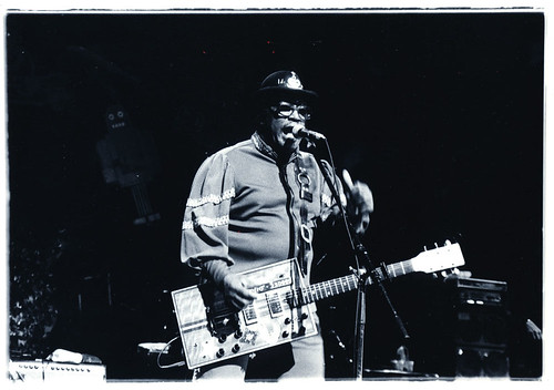 Immagine in bianconero di Bo Diddley, con occhiali e cappello, durante un concerto con una chitarra molto particolare la cui cassa è di forma rettangolare (“bo diddley” by masao nakagami is licensed under CC BY-SA 2.0).
