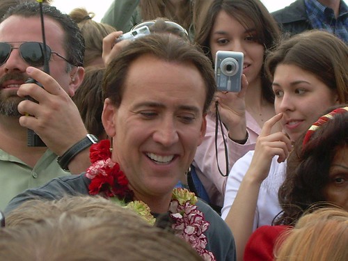 Immagine di Nicolas Cage sorridente in mezzo alla folla con una ghirlanda di fiori al collo (“Nicolas Cage-close up” by Kristin.Standafer is marked with CC BY-SA 2.0).