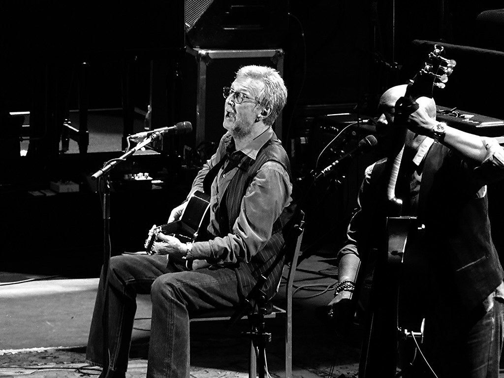 Immagine in bianconero di Eric Clapton, non più ragazzino, ripreso mentre suona la chitarra seduto su una sedia durante un concerto (“Clapton” by Jonathan Bayer is licensed under CC BY-NC-SA 2.0). La musica rock è una filosofia di vita.
