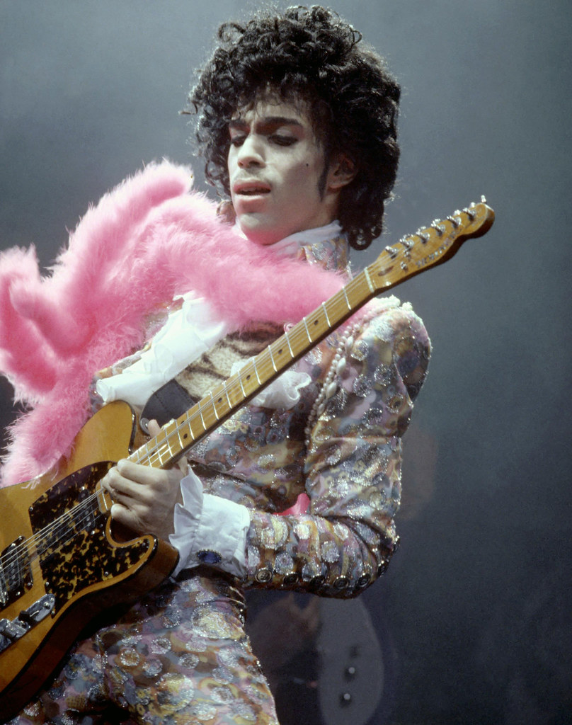 Immagine di Prince mentre suona la chitarra e indossa uno dei suoi abiti "principeschi" ( “Prince-Guitar_pink” by njTare is licensed under CC PDM 1.0).
