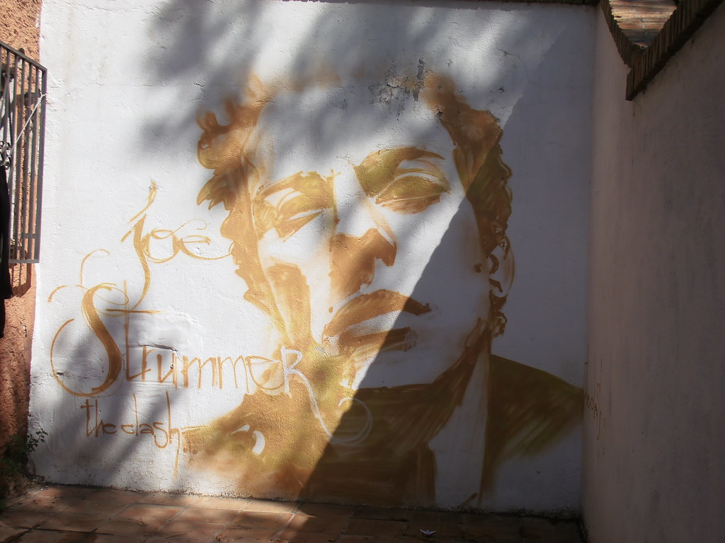 Disegno realizzato sulla parete di un edificio e raffigurante il volto stilizzato di Joe Strummer dei Clash in tonalità giallo oro (“Placeta Joe Strummer, Joe Strummer Square, The Clash, Spain, Andalucia, Granada” by Jeanne Menjoulet is licensed under CC BY-ND 2.0). La musica rock è una filosofia di vita.
