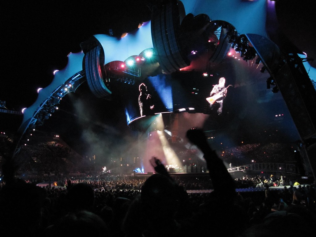 Immagine di un concerto degli U2 da posizione molto distante rispetto al palcoscenico su cui campeggiano grandi schermi che diffondono immagini più ravvicinate della band (“IMG_0176” by elvisripley is licensed under CC BY-NC 2.0). La musica rock è una filosofia di vita.