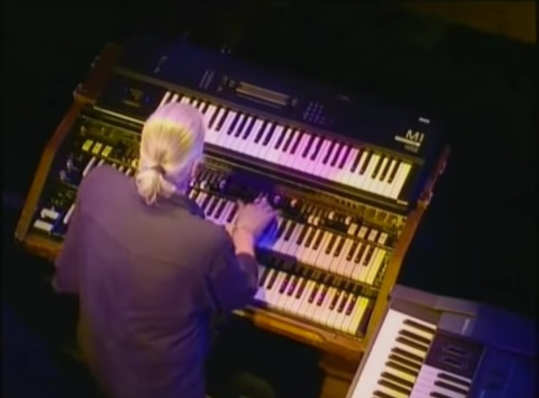 Immagine ripresa dall'alto di un musicista coi i capelli bianchi (raccolti sulla nuca con un codino) mentre suona uno strumento musicale con tastiere multiple.