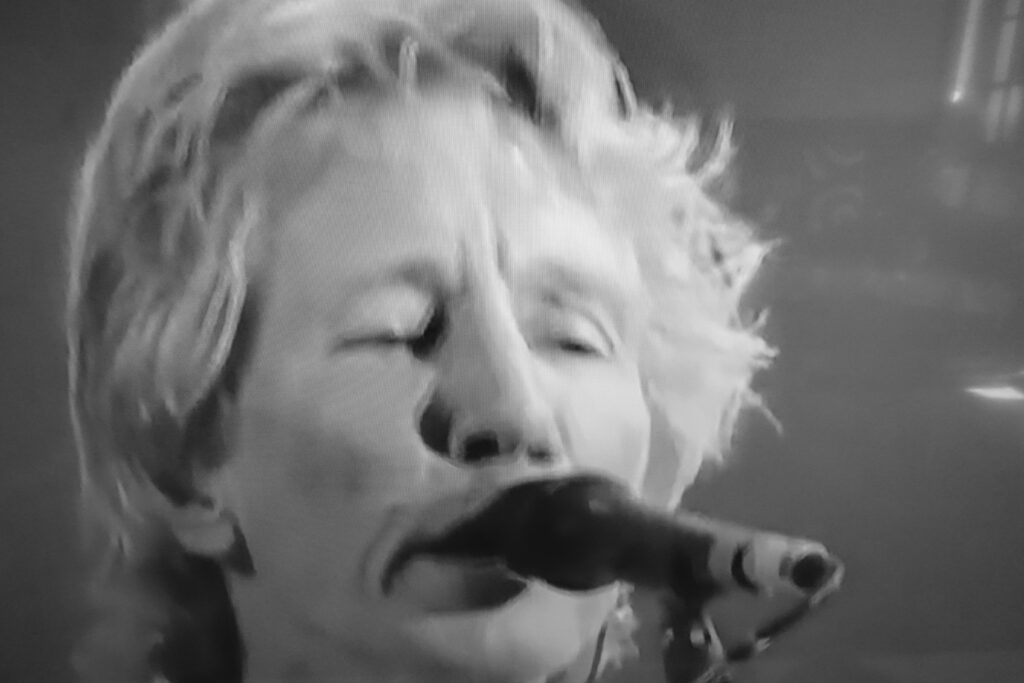 Immagine fotografica in bianconero di Roger Waters in primo piano davanti a un microfono.